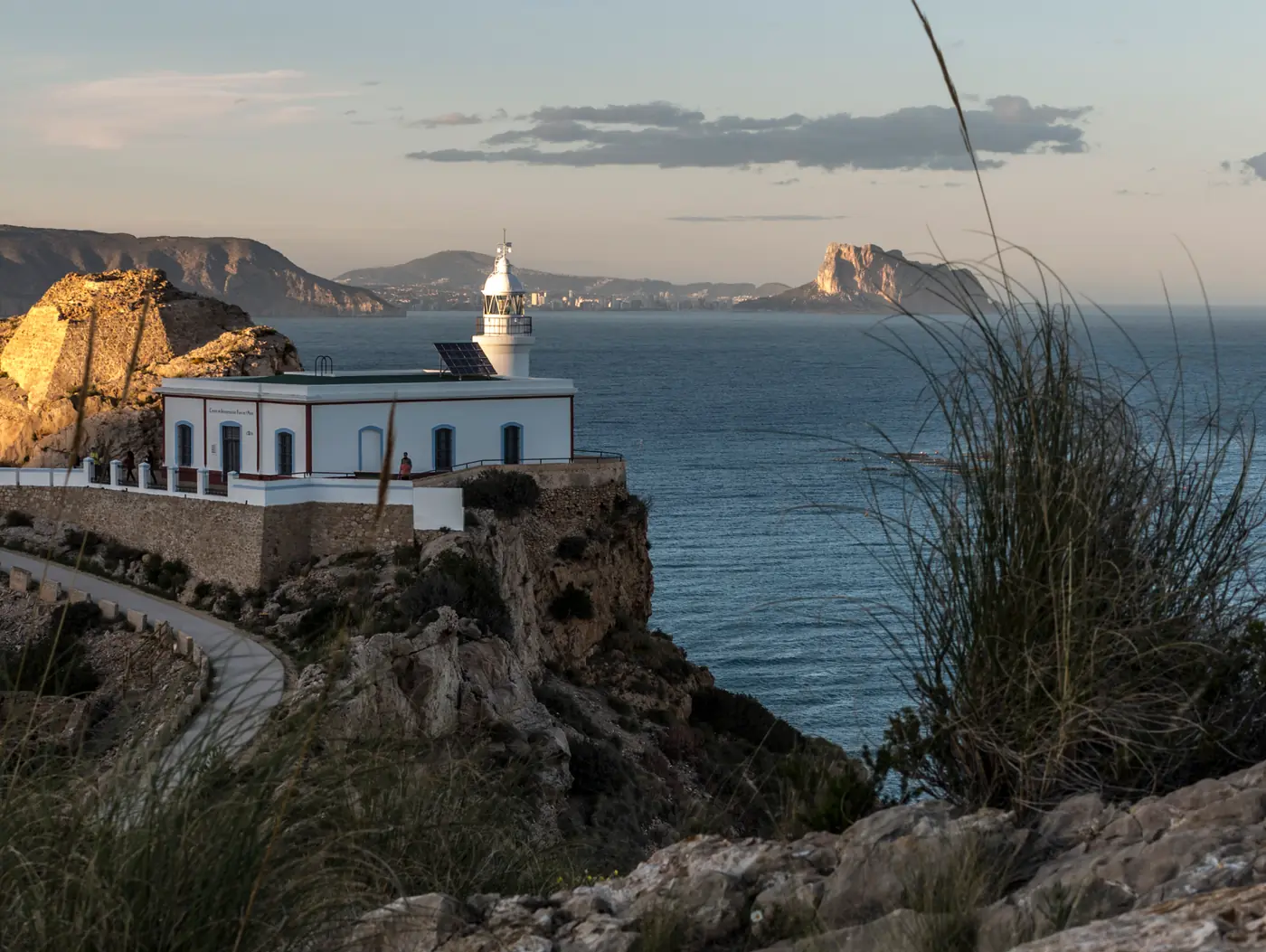 The Albir lighthouse on its clifftop above the Mediterranean at dusk, with the paved access road curving in from the left and the Peñón de Ifach visible across the bay