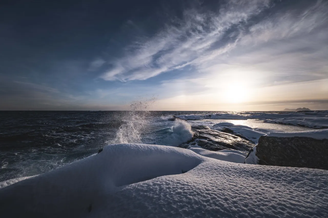 Waves crash against a snow-covered rocky coastline under a dramatic winter sky at sunset.