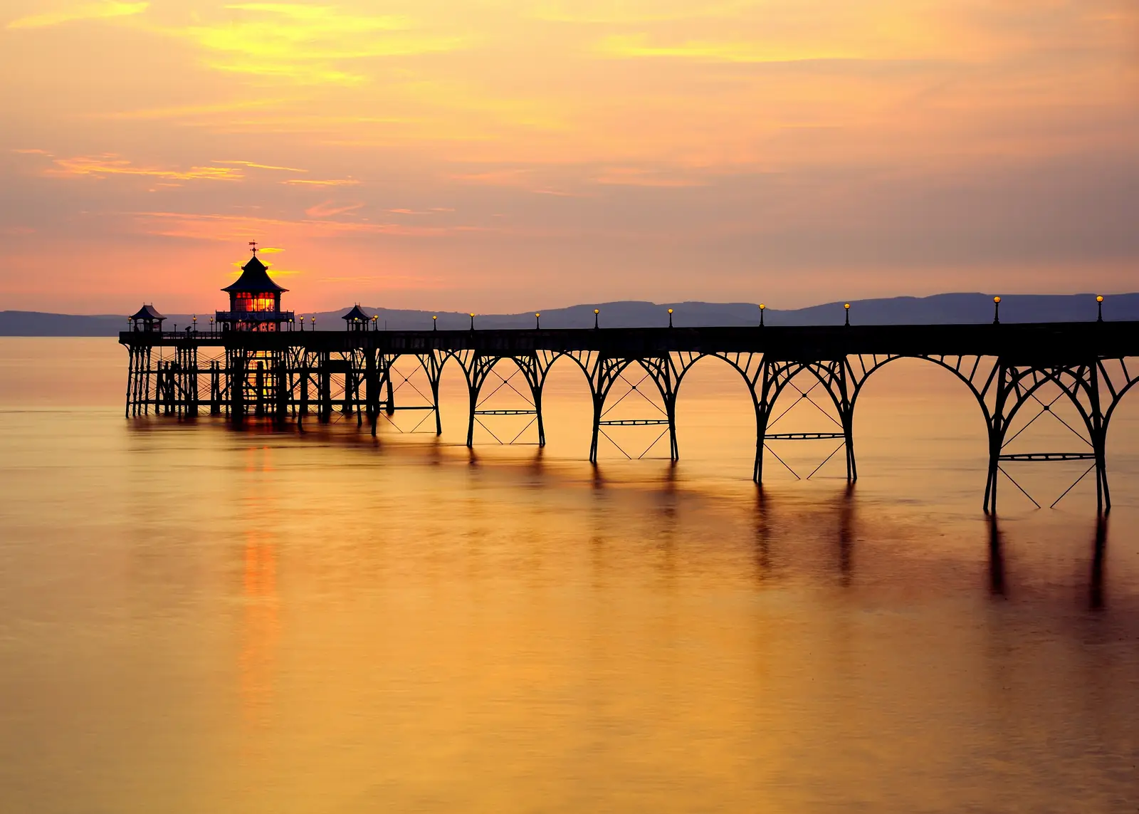 Clevedon Pier silhouetted against an orange sunset over the Severn Estuary, its iron arches reflected in still water