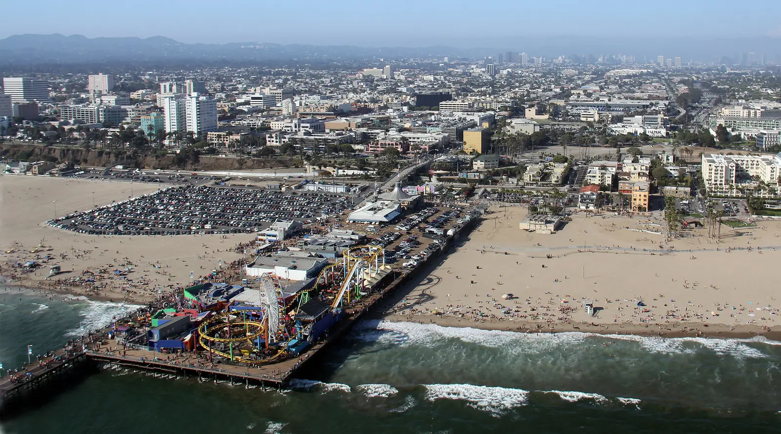 Santa Monica Pier from the air — the wooden pier and its funfair reaching into the Pacific, a wide band of sand either side, and the city stretching inland behind