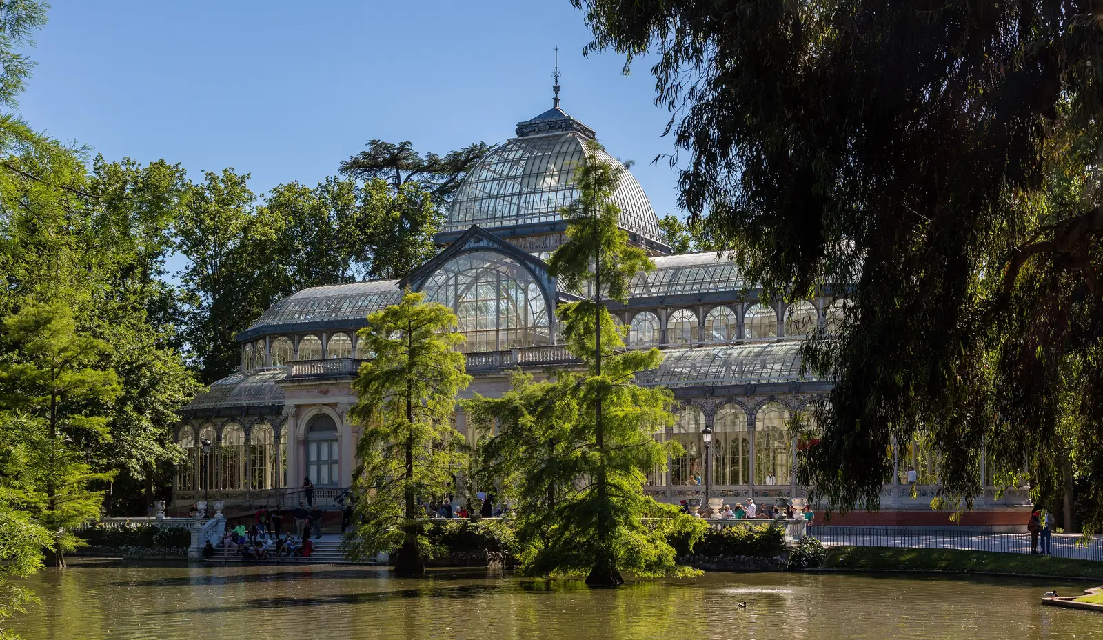 The Palacio de Cristal in Madrid's Parque del Retiro — a glass and iron pavilion reflected in its lake, with the stepped entrance on one side and a paved path around the other