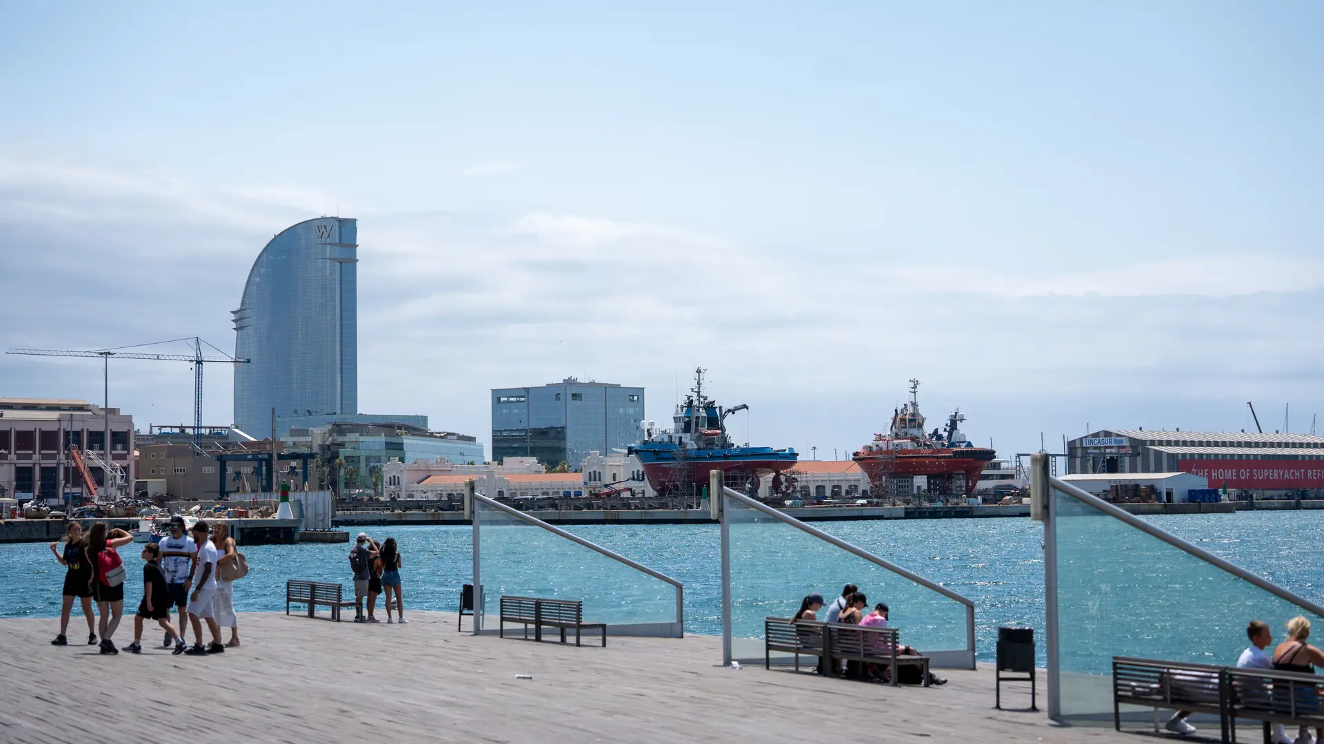 People strolling and sitting on a wide, flat waterfront promenade beside the harbor in Barcelona on a sunny day