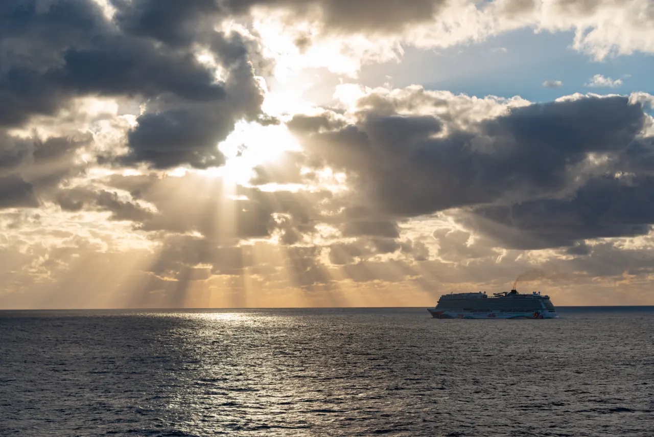 Dramatic sunlight breaks through clouds over a peaceful sea with a cruise ship on the horizon.