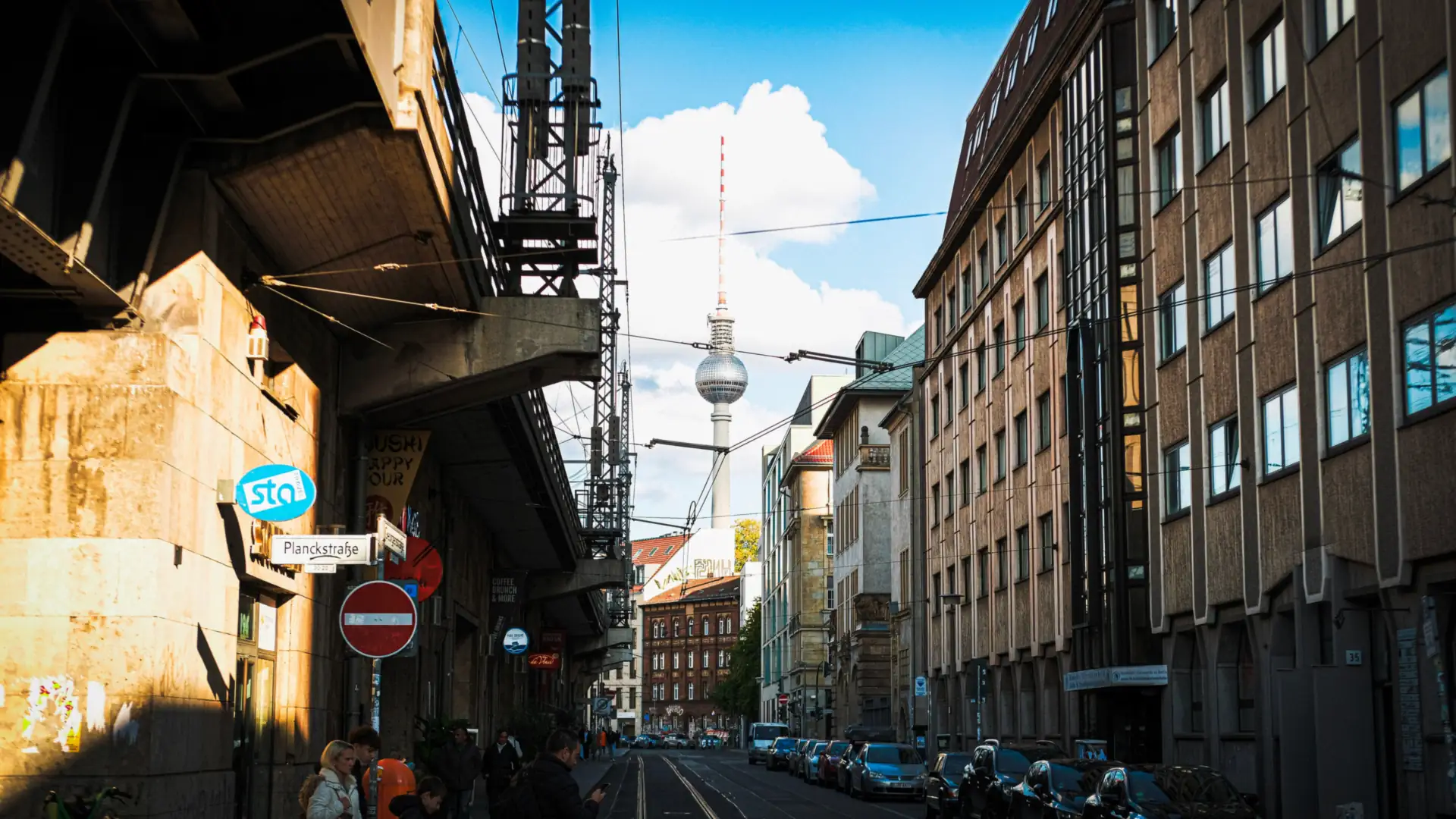 A Berlin street under an elevated railway with apartment blocks and the Fernsehturm TV tower rising against a blue sky