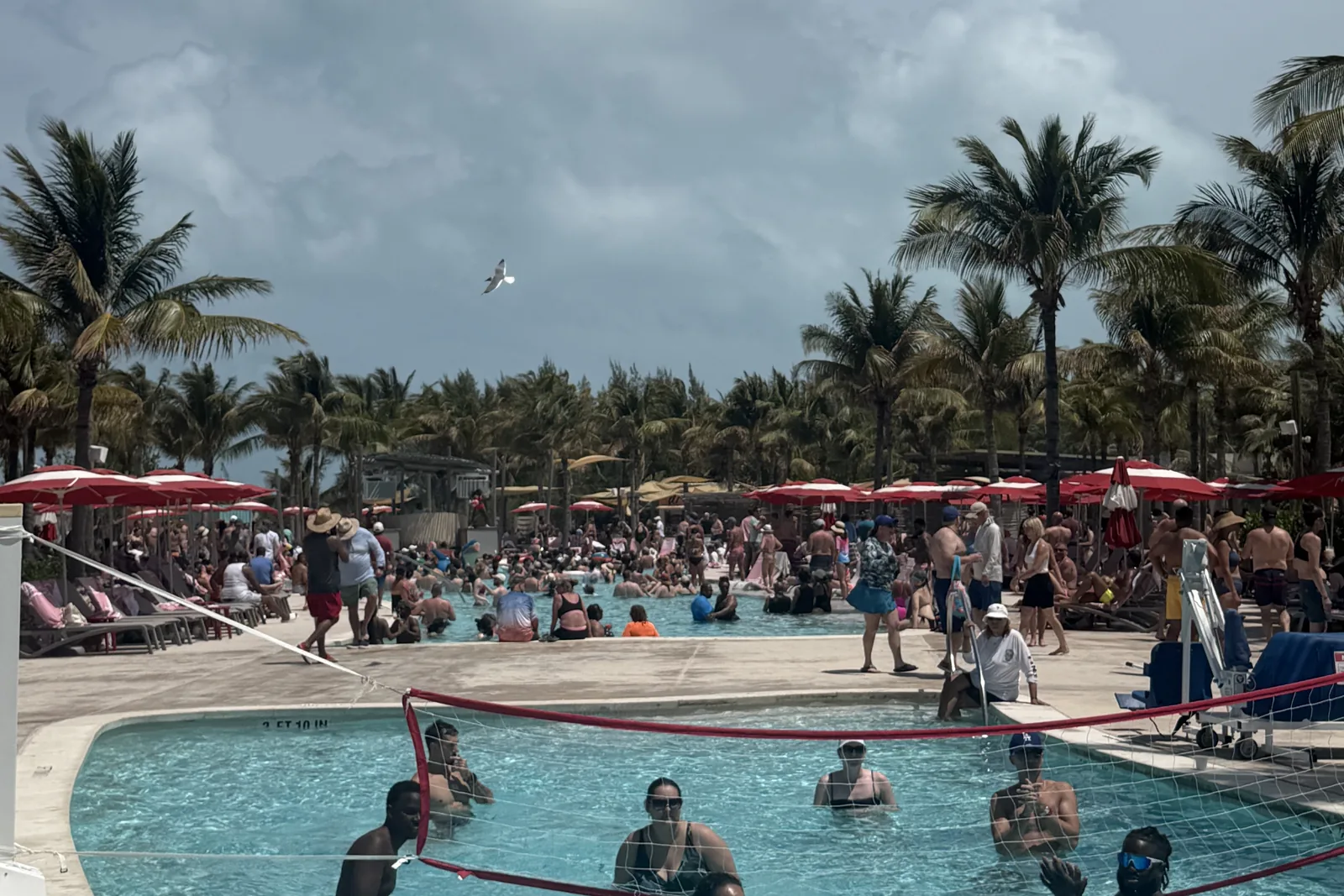 The Beach Club pool crowded with sailors on a call day, red parasols and a volleyball net