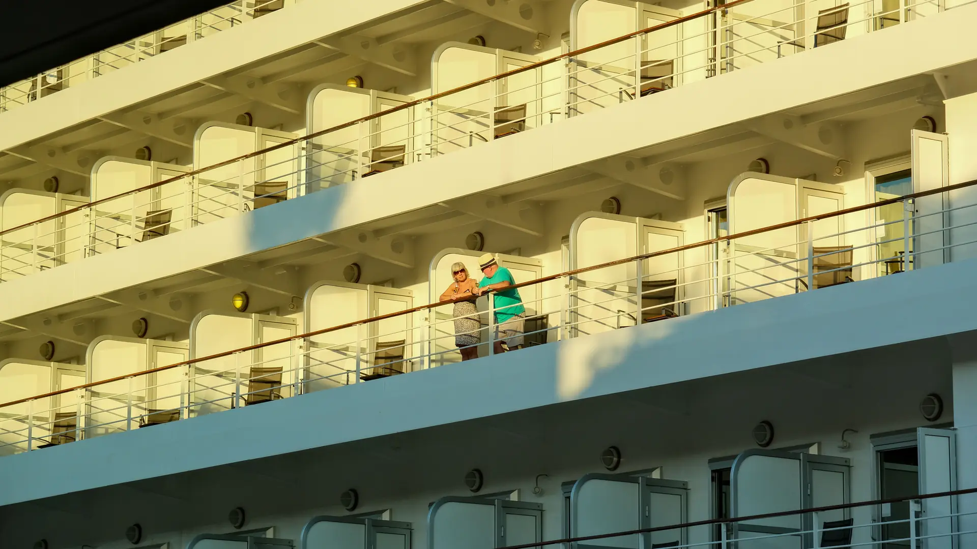 Two adults relaxing on a sunny cruise ship balcony among rows of stateroom balconies