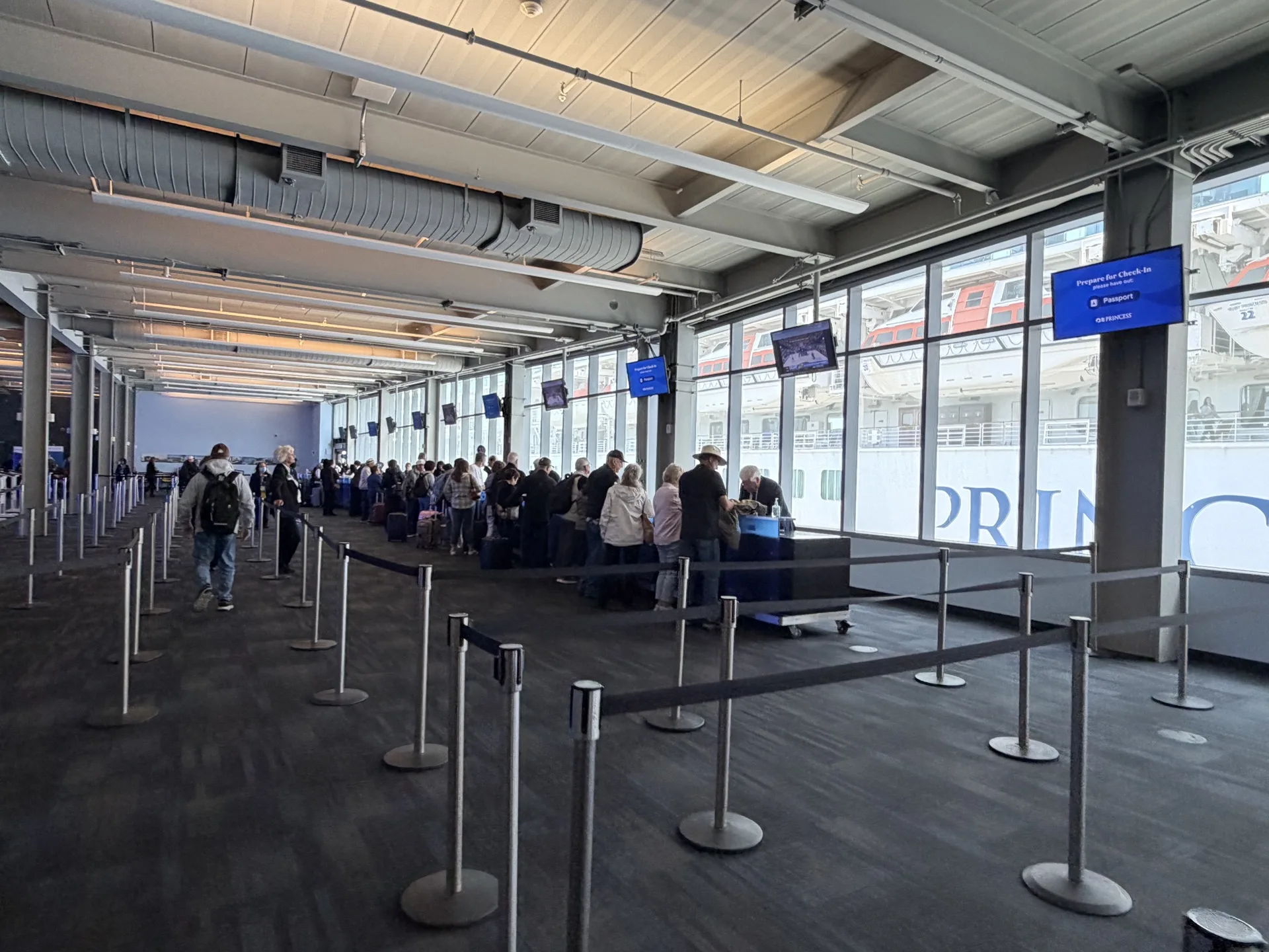 Passengers queuing in a cruise terminal at embarkation, with the ship visible through the terminal glass