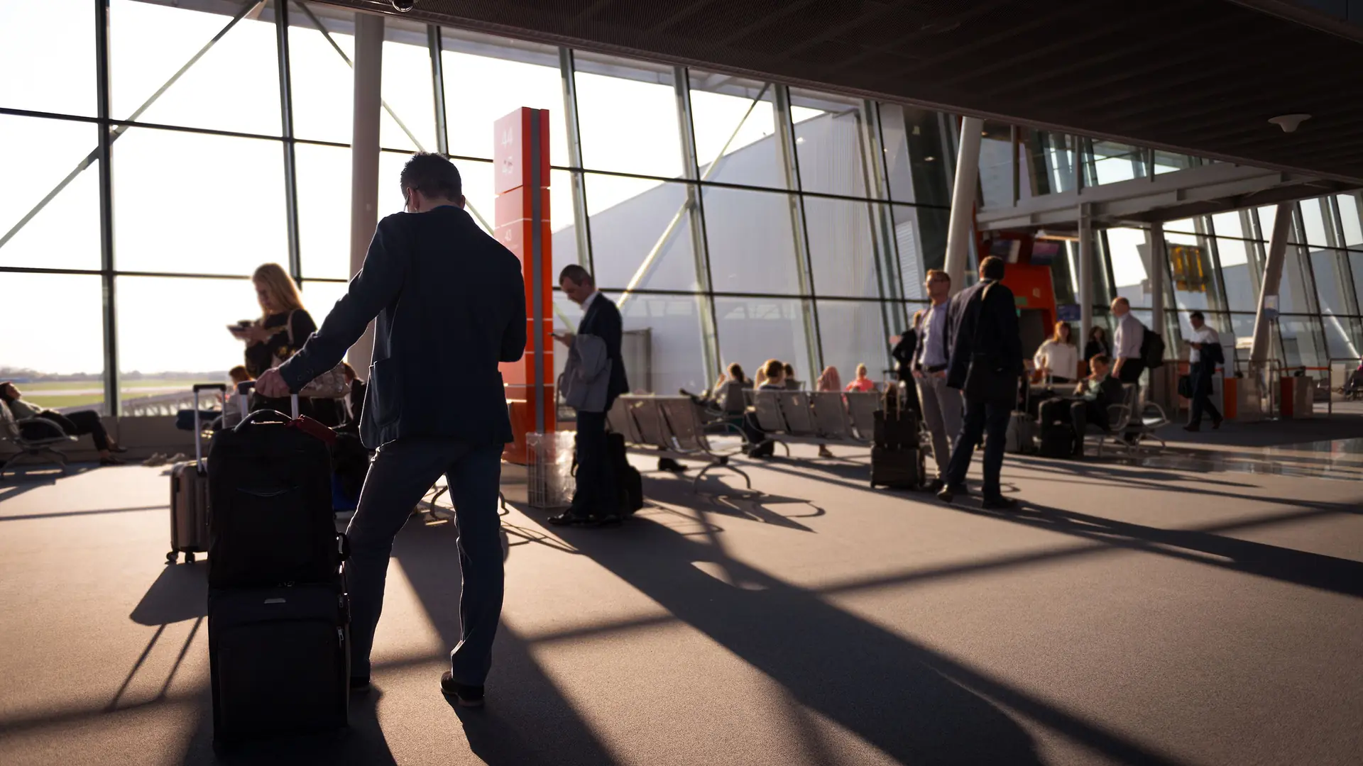 Travelers with rolling carry-on suitcases walking through a bright, sunlit glass-walled terminal