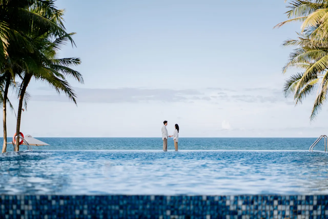 A couple stands by an infinity pool, gazing at the tropical ocean on a sunny day.