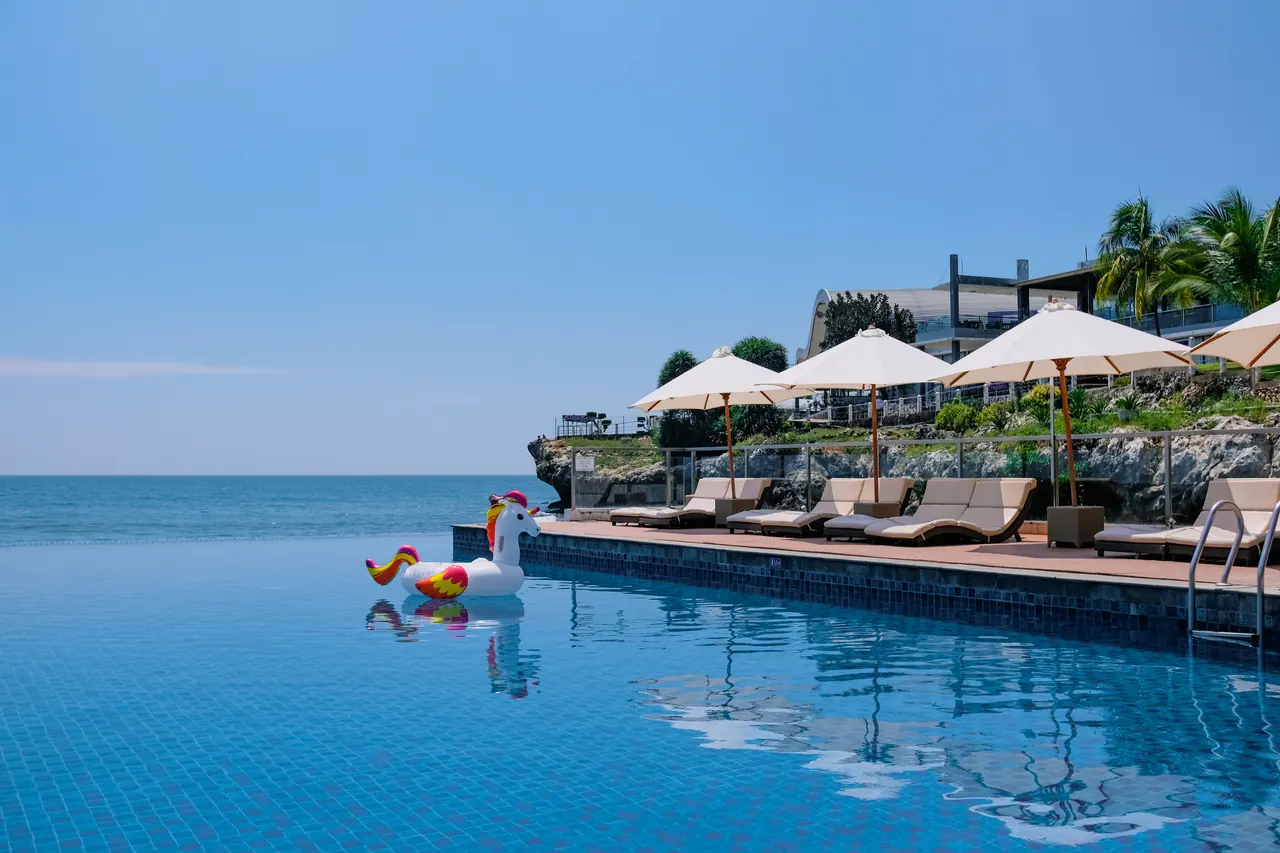 A tranquil beachside infinity pool with sunbeds, umbrellas, and a playful unicorn float under the summer sun.
