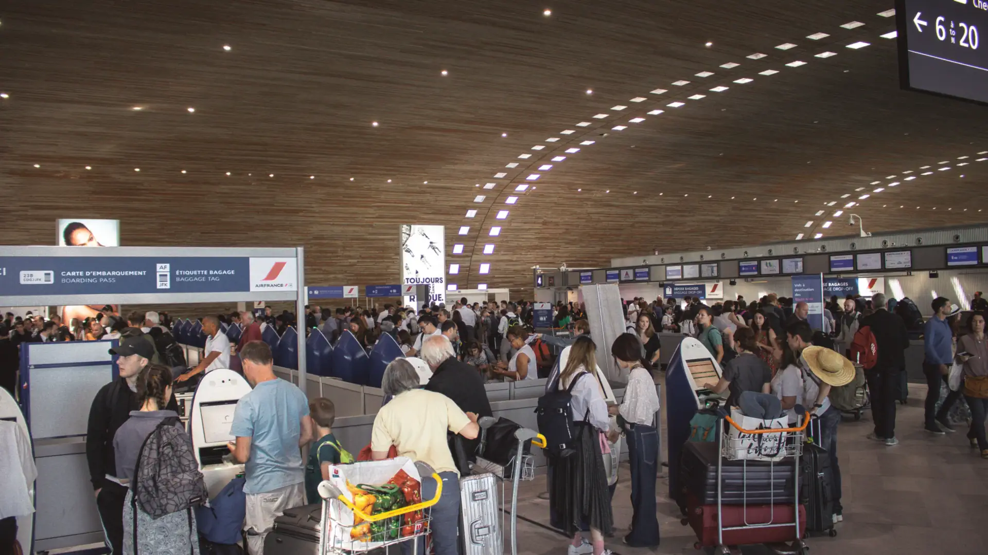 A crowded airport arrivals hall with travelers and luggage carts lined up at border control under a curved wooden ceiling