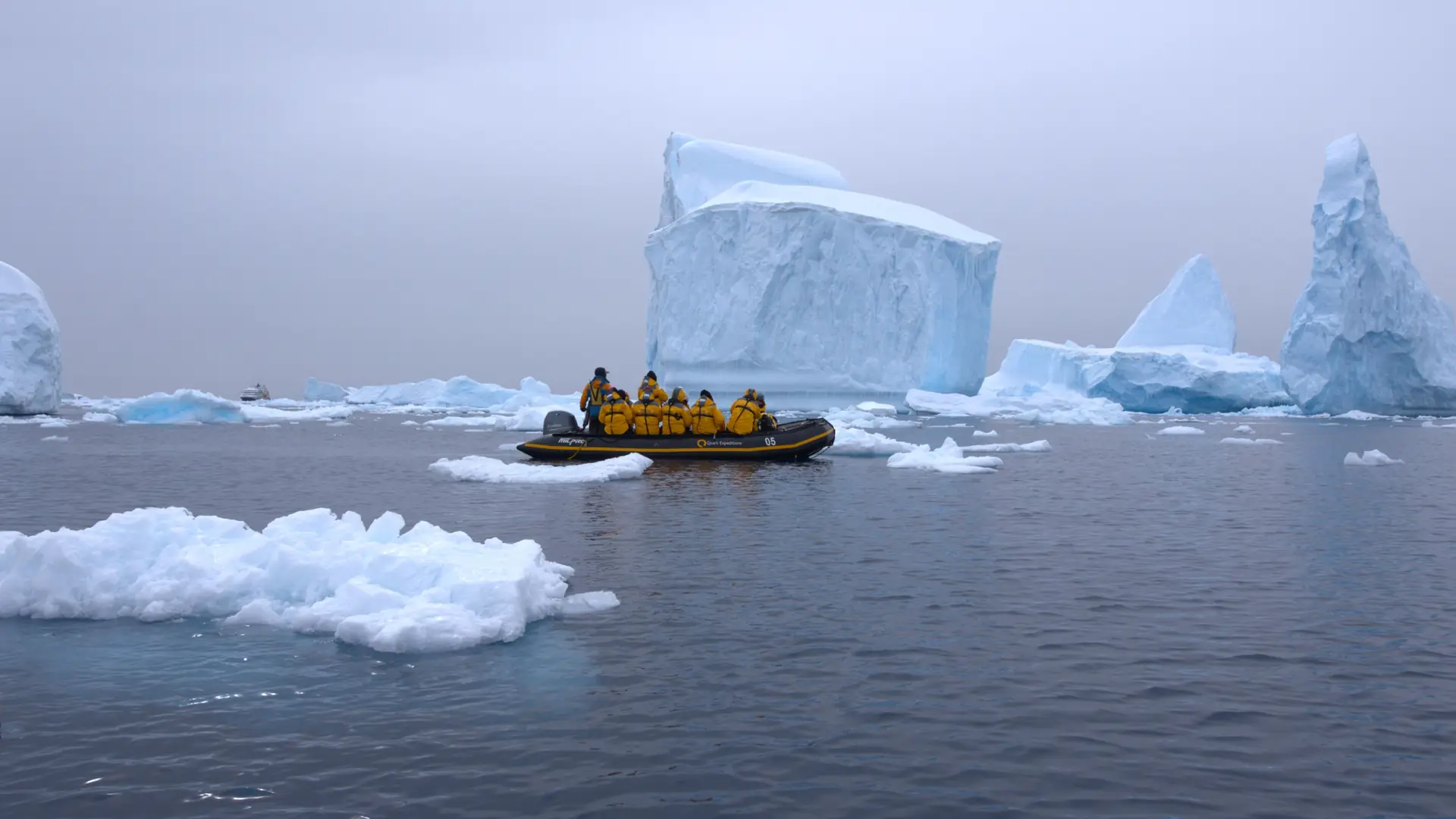 A Zodiac full of passengers in bright expedition jackets cruising grey water past towering white icebergs in Antarctica