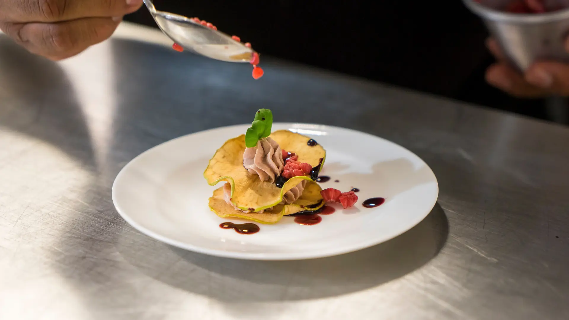 A chef’s hand drizzling berry sauce beside a delicate plated dessert on a white plate in a restaurant kitchen
