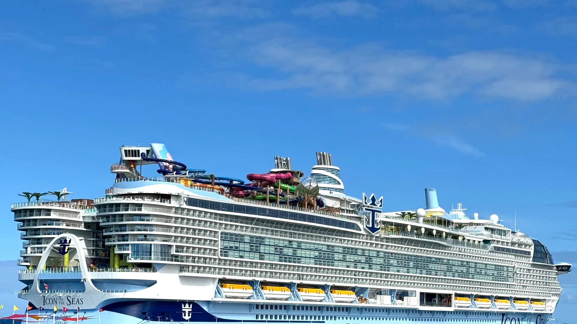 Icon of the Seas alongside at a Caribbean pier under clear sky, her waterslides and upper decks visible along the full length of the hull