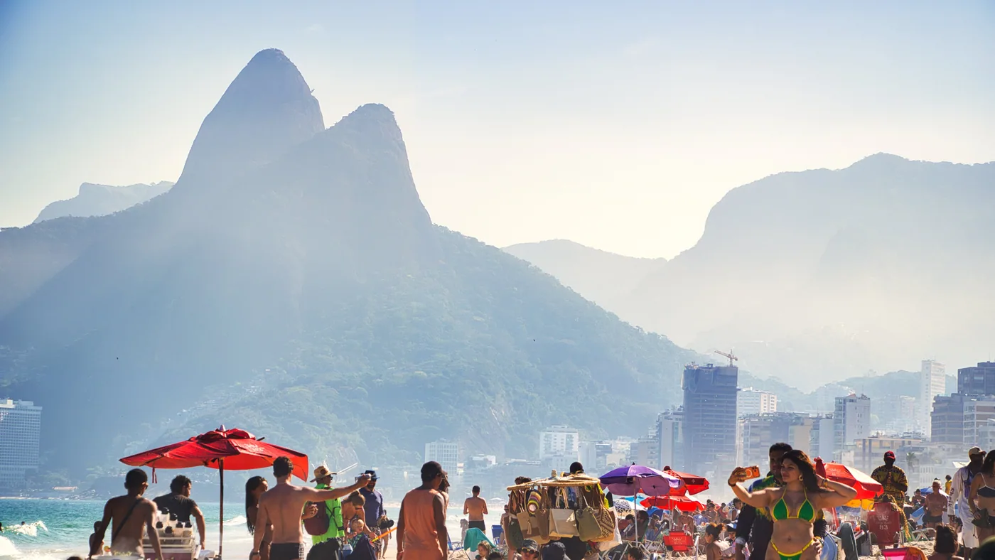 Ipanema Beach crowded on a clear day, with the Dois Irmãos peaks rising at the far end