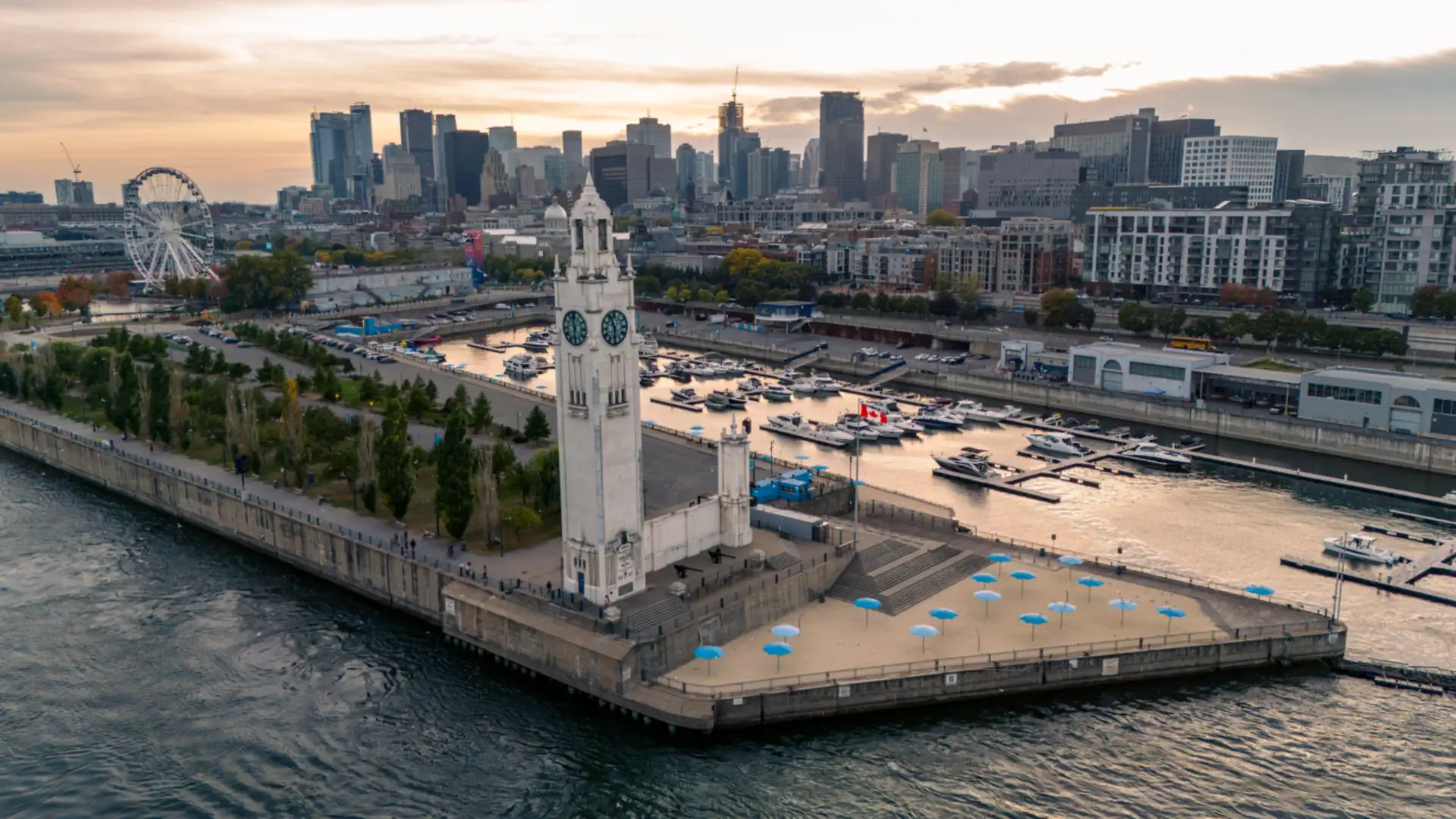 An aerial view of Montréal’s Old Port Clock Tower on a pier beside the river with the downtown skyline behind at sunset