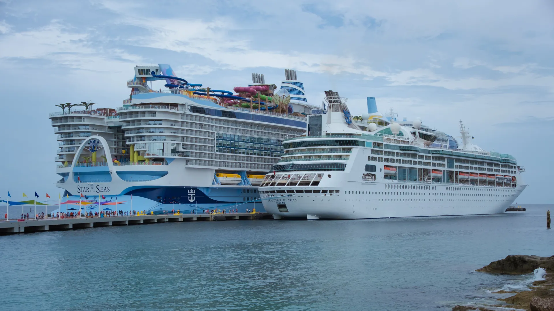 Two cruise ships docked side by side at a busy Bahamian port under a bright sky, with turquoise water in the foreground