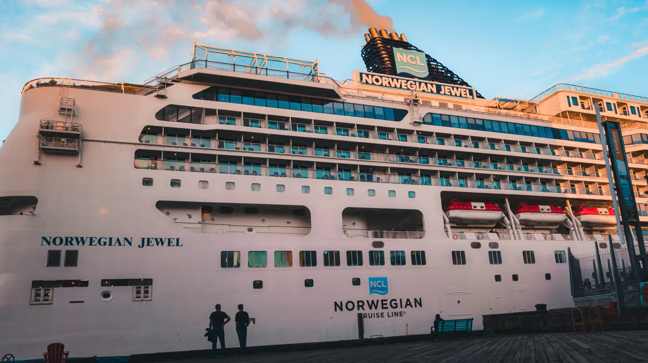 Side view of Norwegian Jewel cruise ship docked, basking in sunset light with passengers on shore.