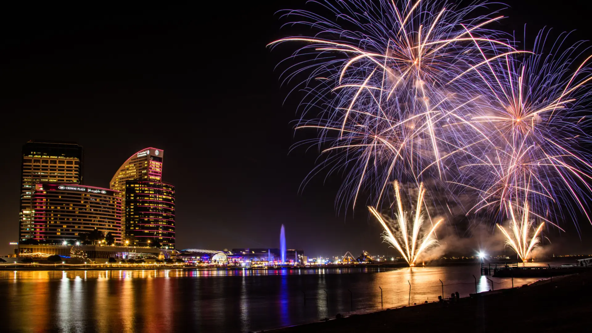 Fireworks bursting in gold and white above a harbour city skyline at night