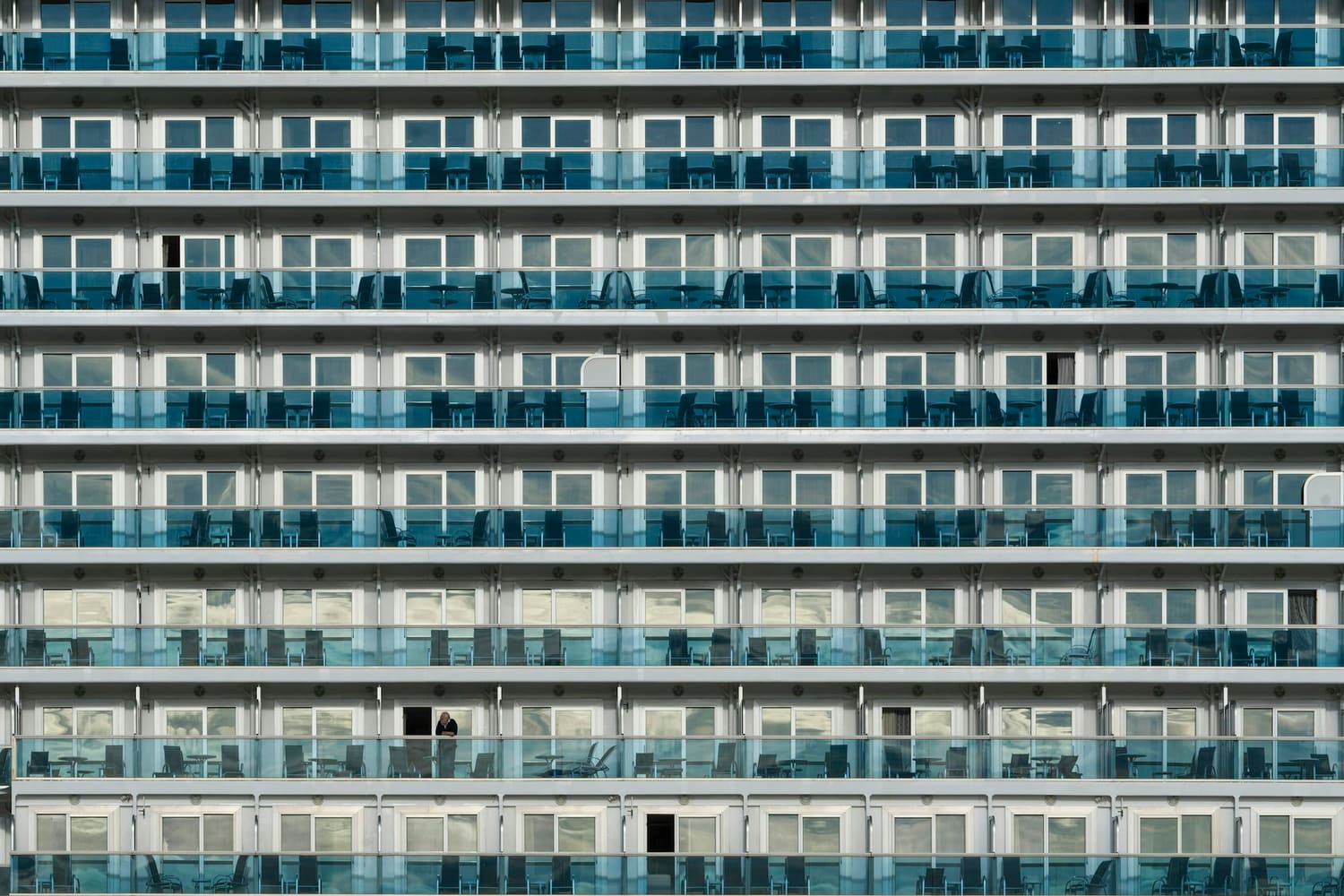 A repeating grid of stacked cruise ship verandas with a single guest on one balcony