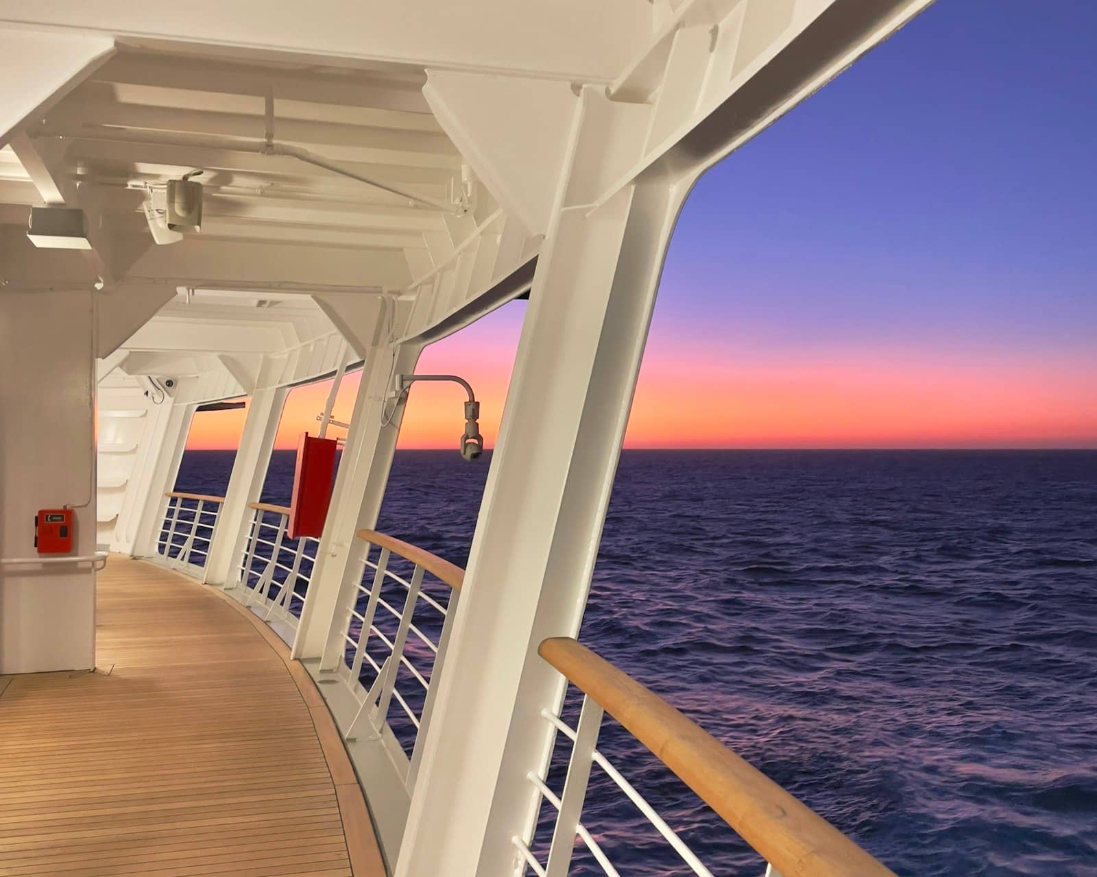 An empty curved teak promenade deck of a cruise ship at sunset, looking out over a calm ocean