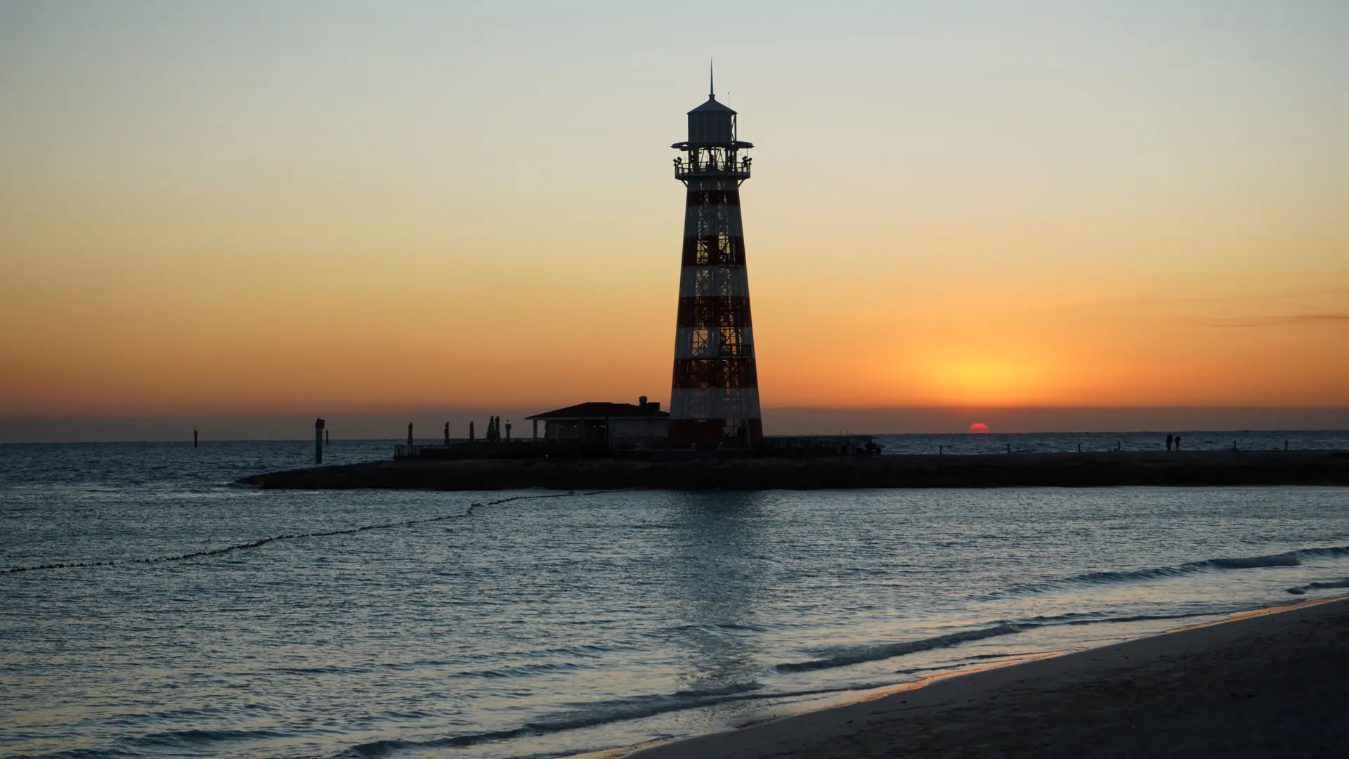 A lighthouse silhouetted against an orange and pink sunset over calm ocean water