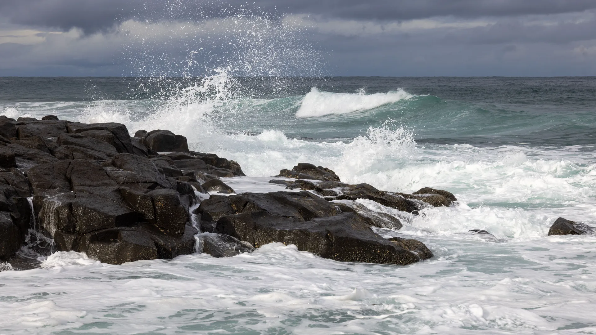 Heavy ocean swell breaking white against a rocky shoreline under a low grey sky