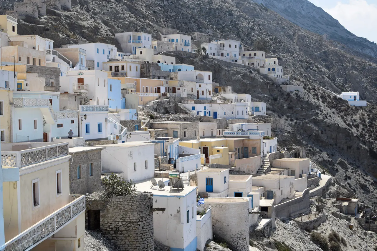 Charming Greek island village with whitewashed houses nestled into a rocky hillside, under a bright sky.