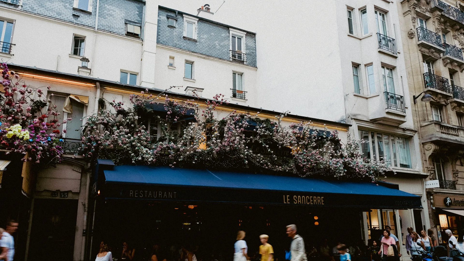 A busy Paris street with a restaurant terrace and a flower-covered facade