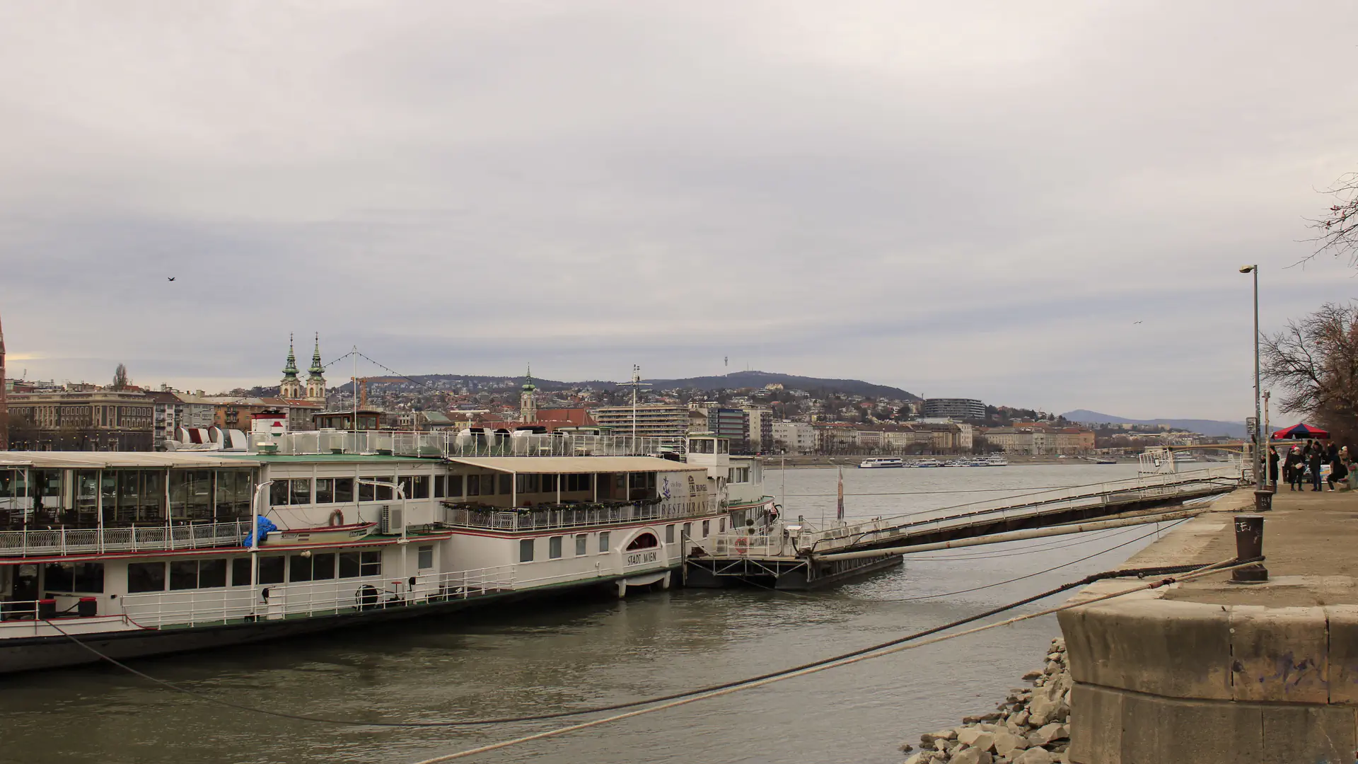 A river cruise vessel moored against a stone embankment on the Danube in Budapest, with a narrow gangway running to the quay