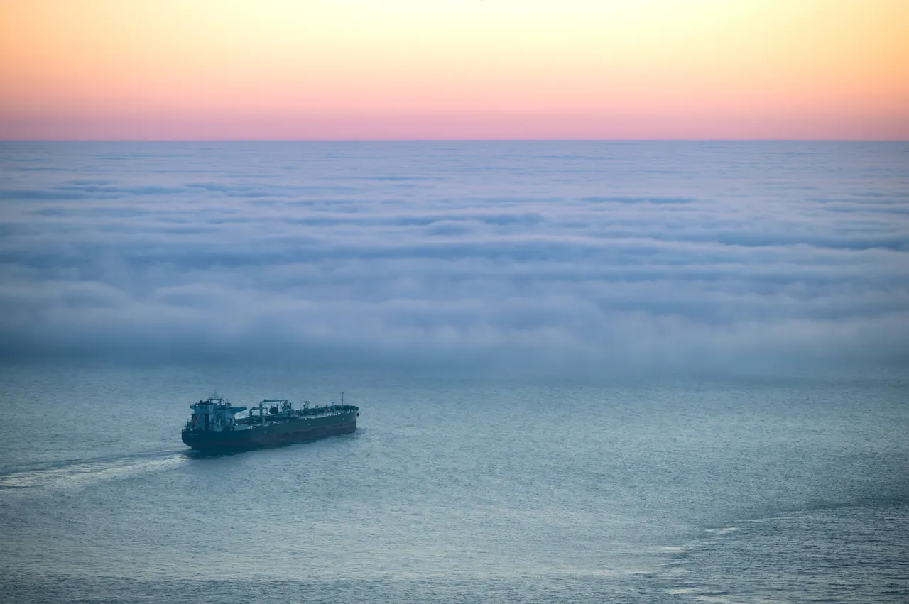A solitary ship sails through calm ocean waters beneath a vibrant, foggy sunset horizon.