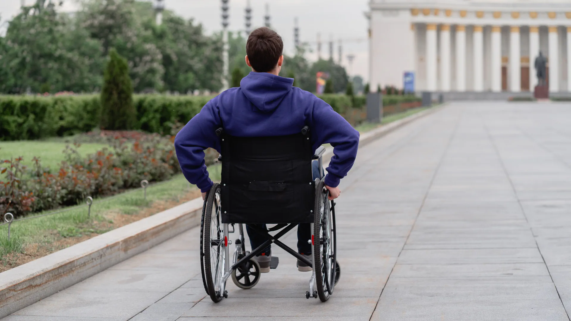 A man in a wheelchair, seen from behind, propelling himself along a wide paved promenade in a park
