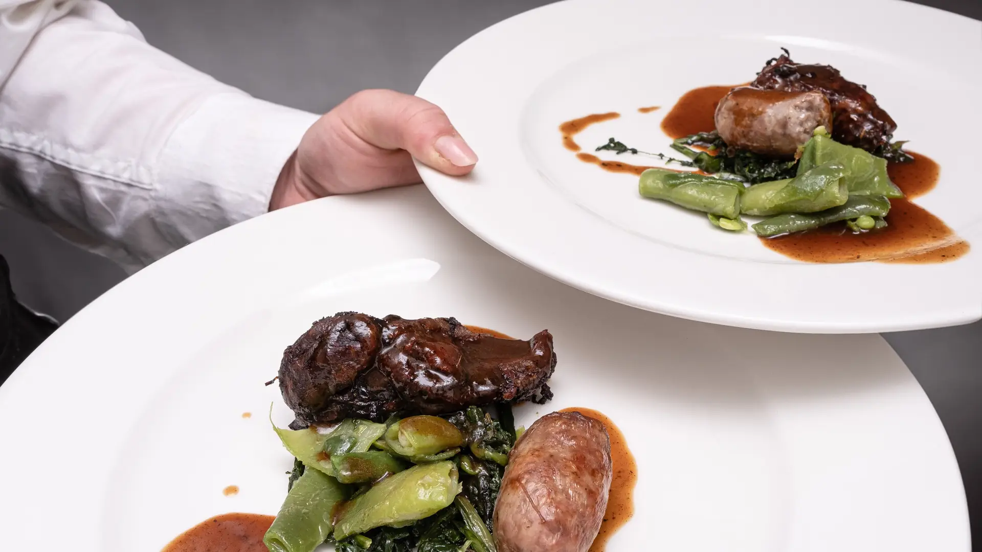 A server’s hand presenting two white plates of braised meat, sausage and greens in a rich sauce