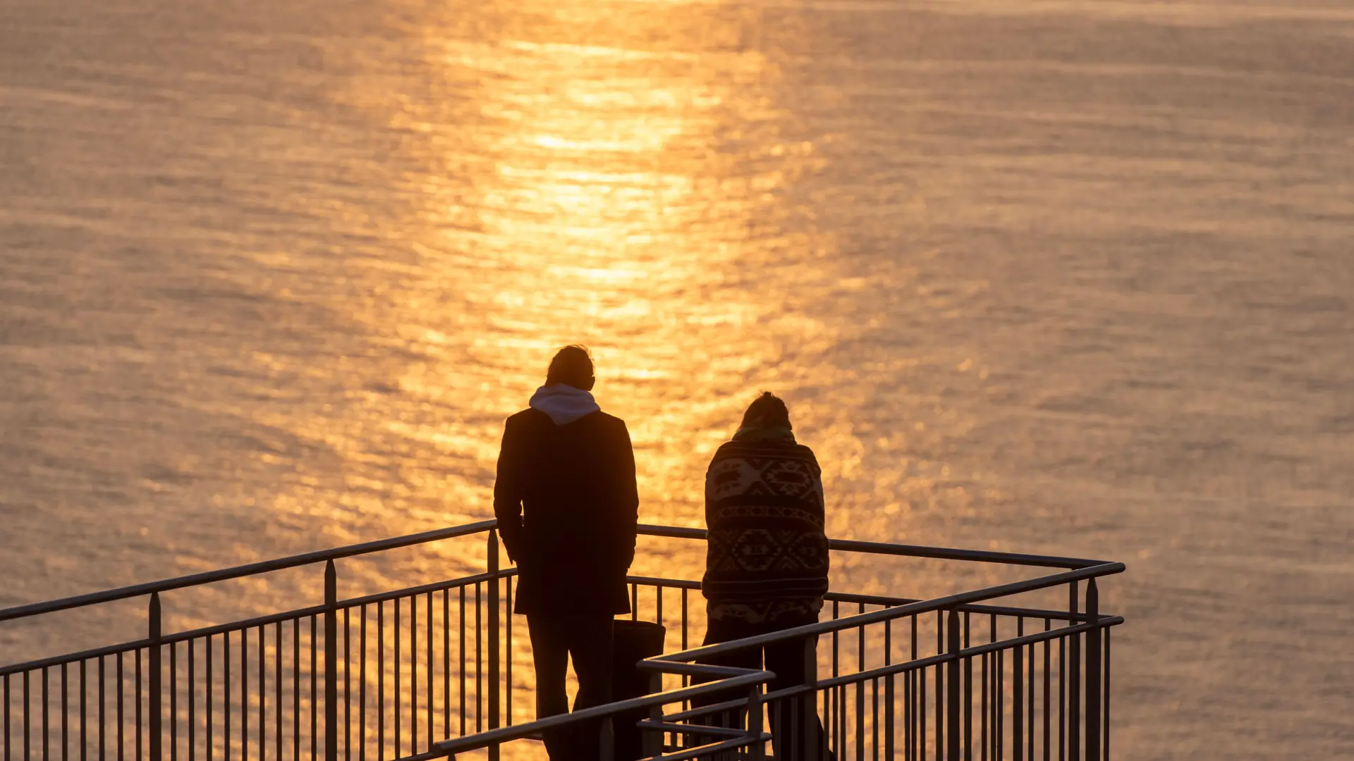 Two people standing at a ship railing silhouetted against a golden sunset over the sea