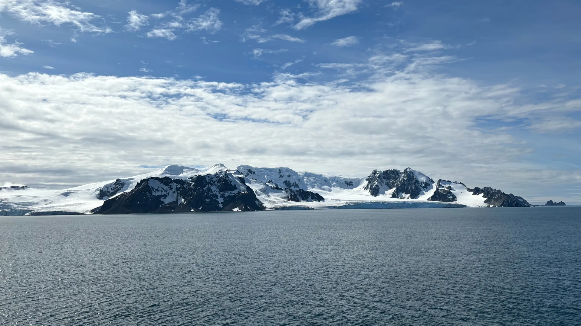 Snow-covered mountains and glaciers along the Antarctic coastline under a blue sky with drifting cloud, seen across calm gray water