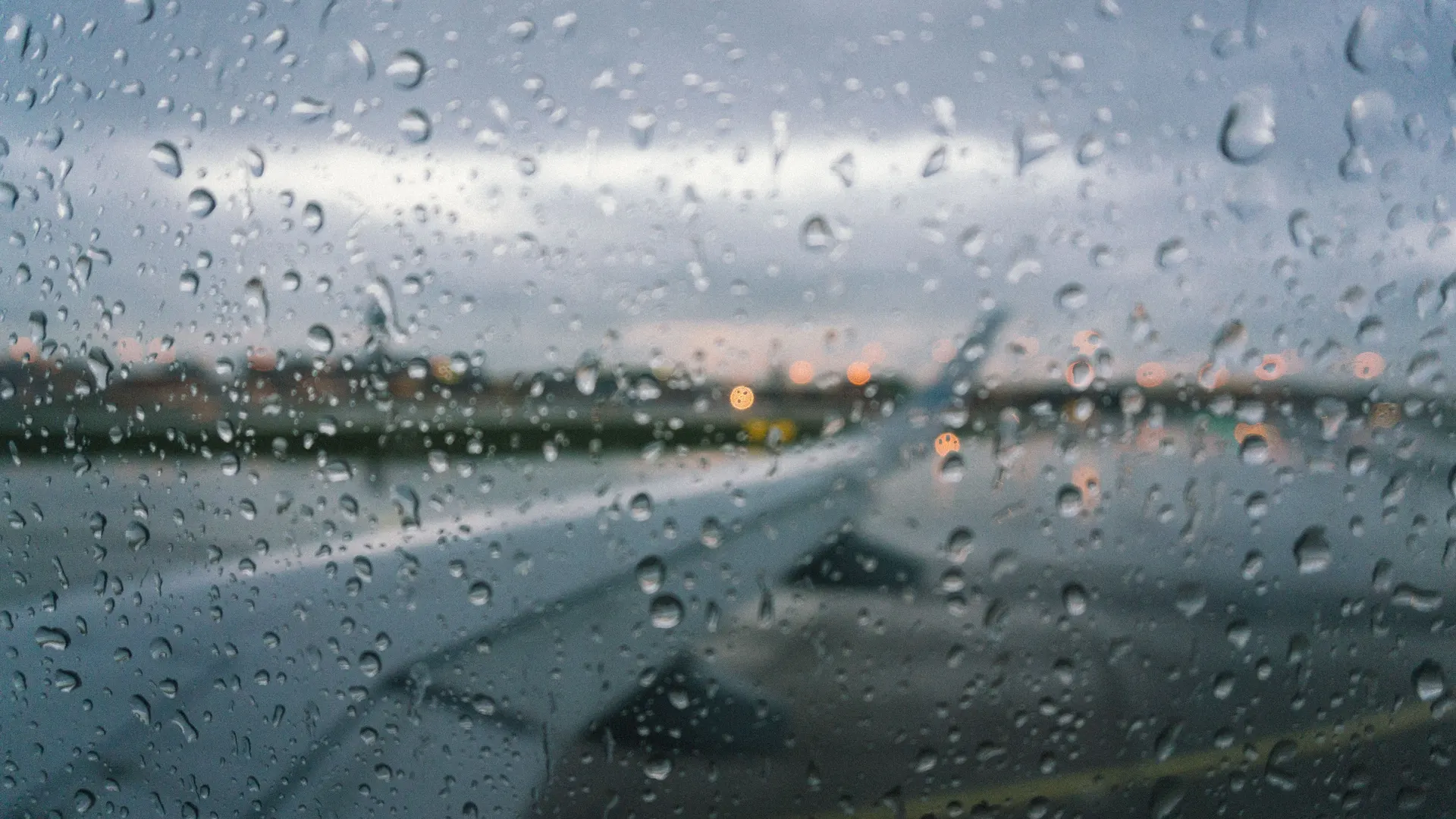 Raindrops on an airplane window, with the wing and a wet runway beyond