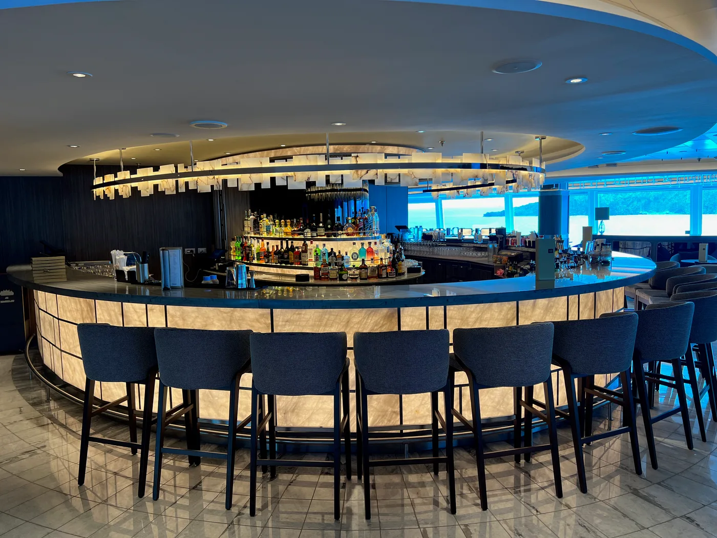 A curved, softly lit cruise ship lounge bar with a full back-bar of spirits bottles and sea views beyond