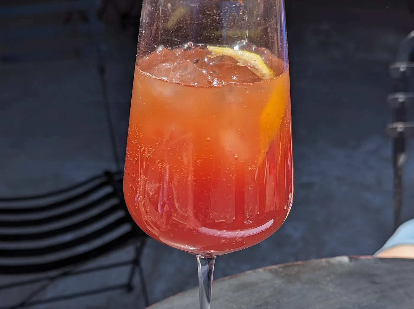 A tall stemmed glass of a red-orange sparkling zero-proof spritz with citrus, on a slate table